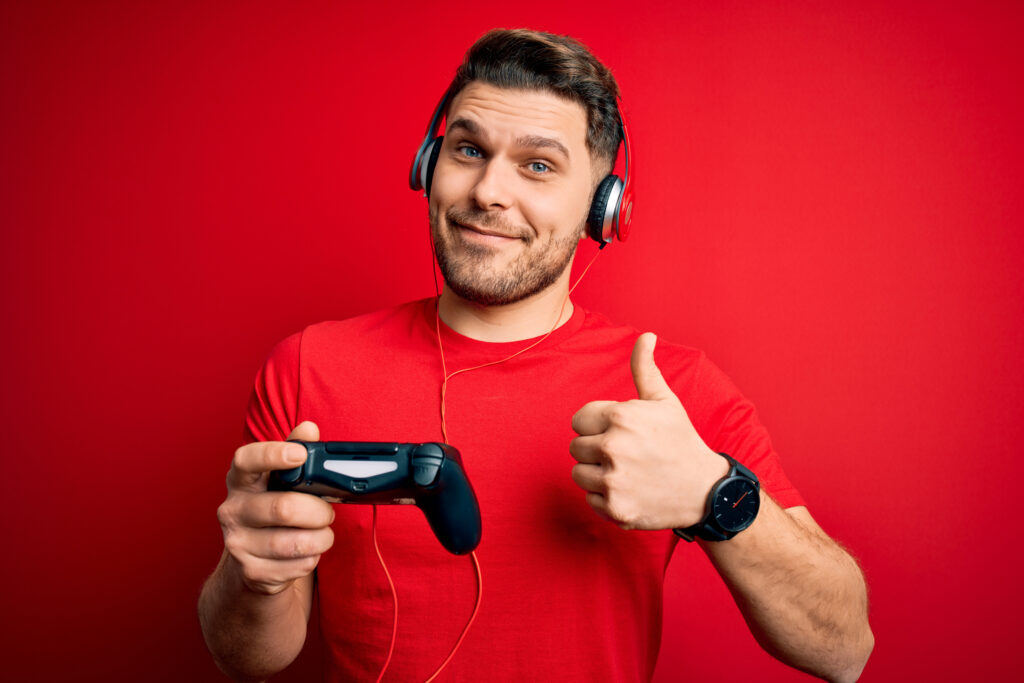 Gamer in a red shirt and red Background holding a game controller in one hand while giving thumbs up with the other.